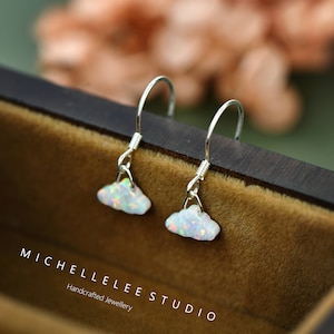May include: A pair of silver hook earrings featuring small, white, iridescent cloud-shaped charms with flashes of orange and pink. The text MICHELLELEE STUDIO Handcrafted Jewellery is printed in white on the dark brown surface below the earrings.