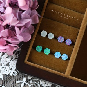 May include: A close-up of six pairs of hexagonal opal earrings in a wooden box. The earrings are in various colors, including white, blue, green, and purple. The box has a label that reads "MICHELLELEE STUDIO Handmade Jewellery".