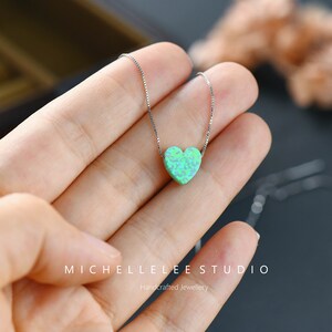 K&ouml;nnte beinhalten: Eine filigrane Silberkette mit einem herzf&ouml;rmigen Anh&auml;nger in Gr&uuml;n- und Blaut&ouml;nen, der wie Opal aussieht. Die Kette wird in einer Hand pr&auml;sentiert. Der Markenname "MICHELLELEE STUDIO" und der Text "Handcrafted Jewellery" sind sichtbar.