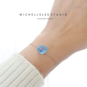 May include: A delicate silver chain bracelet featuring a single, large, light blue heart-shaped bead. The bracelet is worn on a woman's wrist.