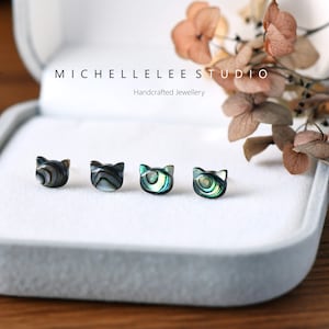 May include: Four small cat-shaped earrings made of iridescent abalone shell. The earrings are displayed in a white velvet jewellery box.