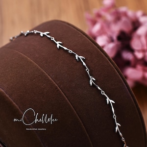 Puede incluir: Una delicada pulsera de plata con un diseño de hojas repetidas. La pulsera se muestra sobre una superficie marrón, con un arreglo floral rosa borroso en el fondo. La pulsera es una pieza de joyería artesanal.