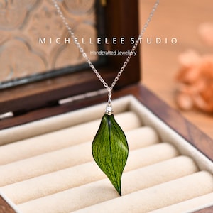 May include: A silver necklace featuring a green leaf-shaped pendant with detailed veining and a small, clear gem. The necklace is presented in a wooden jewellery box. The text "MICHELLELEE STUDIO Handcrafted Jewellery" is visible.