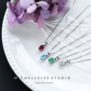 May include: Five silver necklaces with different coloured gemstones. The necklaces are on a white plate with a floral background. The gemstones are red, blue, green, and two light blue. The text "MICHELLE LEE STUDIO" and "Handcrafted Jewellery" is at the bottom of the image.
