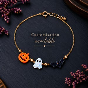 May include: A gold-tone chain bracelet featuring three charms: an orange pumpkin, a white ghost, and a dark purple bat. The bracelet includes small gold beads between the charms and a lobster clasp. The text Customisation available is centred.