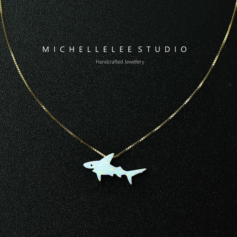 May include: A gold chain necklace with a white opal shark pendant. The pendant is a detailed silhouette of a shark with a white opal stone filling the shape. The necklace is on a black background. "MICHELLELEESTUDIO Handcrafted Jewellery" is written in white text at the top of the image.