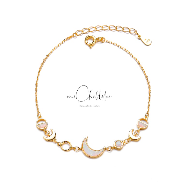 May include: A gold-colored bracelet featuring a crescent moon and circular opal charms. The bracelet has a delicate chain and a lobster clasp closure. The text "mi Chellelee" is visible in the image.
