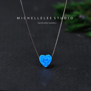 May include: A silver necklace featuring a blue opal heart pendant. The pendant is suspended from a delicate chain. The image is set against a dark background. The text "MICHELLELEE STUDIO Handcrafted Jewellery" is visible.