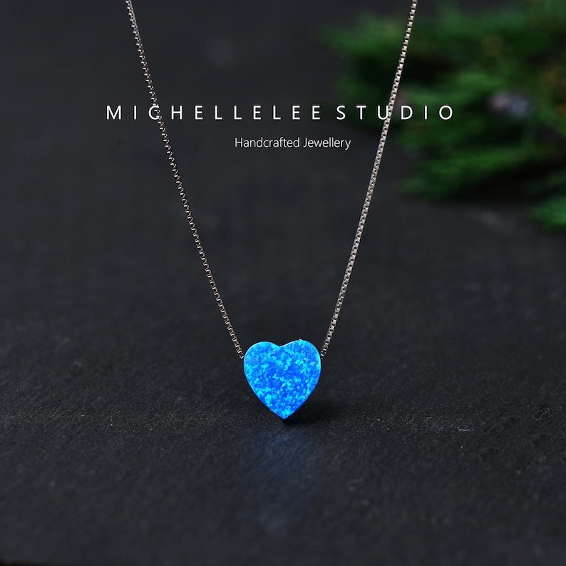 May include: A silver necklace featuring a blue opal heart pendant. The pendant is suspended from a delicate chain. The image is set against a dark background. The text "MICHELLELEE STUDIO Handcrafted Jewellery" is visible.