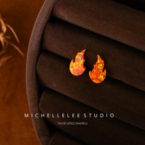 Può includere: Un paio di orecchini a forma di fiamma con una finitura opalescente rosso e arancione. Gli orecchini sono esposti su uno sfondo di velluto marrone. Michelle Lee Studio Gioielli artigianali.