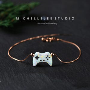 Peut inclure: Un bracelet délicat en chaîne or rose avec un pendentif de manette de jeu vidéo en opale blanche. Le pendentif est détaillé avec des boutons noirs et une croix.