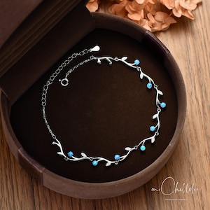 Op de afbeelding: Een zilveren armband met een delicaat wijnstokontwerp, met kleine blauwe edelstenen en bladaccenten. De armband wordt getoond in een bruine sieradendoos, tegen een houten achtergrond.