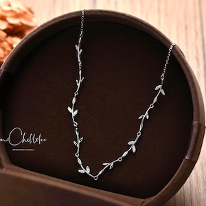 Op de afbeelding: Een delicate zilveren ketting met een bladeren ontwerp, gepresenteerd in een bruine sieradendoos. De ketting heeft kleine, gedetailleerde bladeren en ligt op een houten oppervlak. De merknaam "mi Chellelee" is zichtbaar.