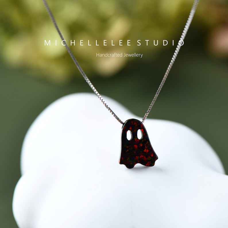 May include: A silver chain necklace with a red and black opal ghost pendant. The pendant has two small holes for eyes. The necklace is on a white surface with a green background.