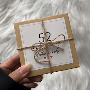 May include: A small, square, brown cardboard box tied with twine. The box has a white label with the number "52" and the words "date cards" in script, plus two small red heart symbols. The box is held by a person.