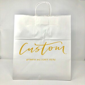 May include: White paper shopping bag with twisted paper handles. The bag features the word "Custom" in gold script, with "OPTIONAL MESSAGE HERE" printed below. The bag is designed for retail or gift use.