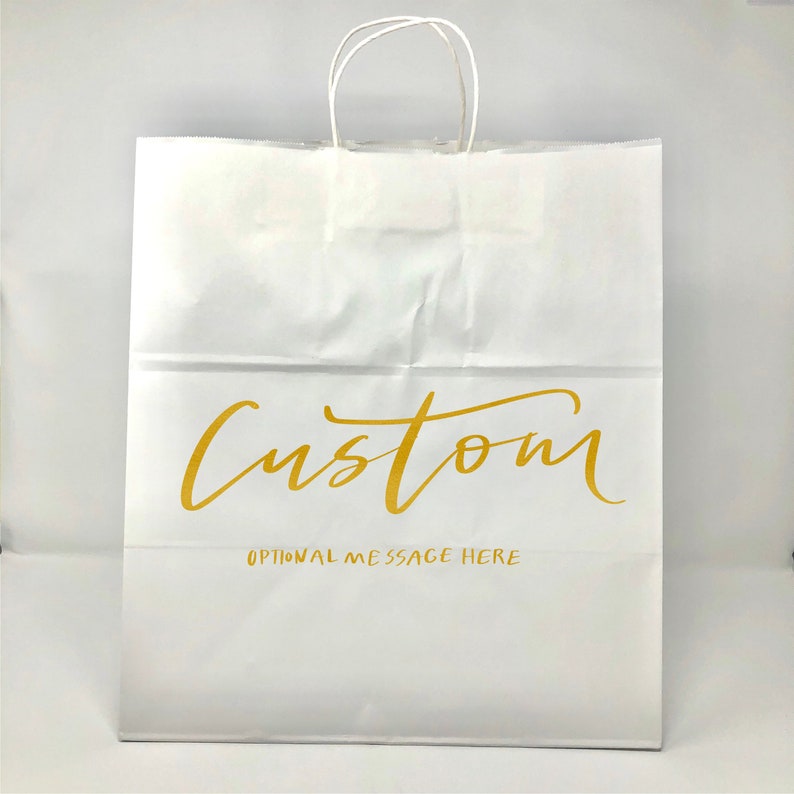 May include: White paper shopping bag with twisted paper handles. The bag features the word "Custom" in gold script, with "OPTIONAL MESSAGE HERE" printed below. The bag is designed for retail or gift use.