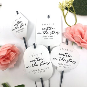 May include: White paper tags with the text "LOVE IS written in the stars" in black script. Each tag has a different shape, including a diamond, rectangle, oval, and a decorative shape. The tags are attached to dark gray sticks.