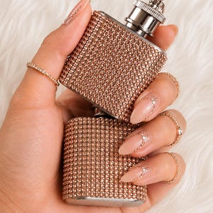 Op de afbeelding: Een rechthoekige metalen heupfles bedekt met kleine, roségoudkleurige strass-steentjes. De fles is voorzien van een zilverkleurige schroefdop met een klein metalen lusje aan de bovenkant voor een veilige afsluiting van de inhoud.