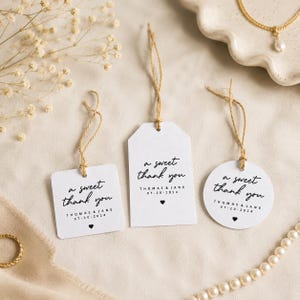 May include: Three white paper gift tags in square, rectangular, and circular shapes, each featuring black printed text that reads a sweet thank you, Thomas & Jane, 20-07-2024, and a small heart icon, all attached with thin brown twine strings.
