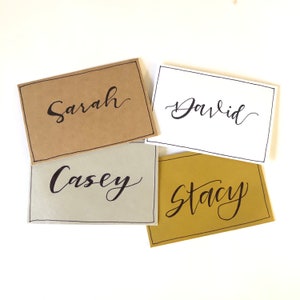 May include: Four place cards with names written in calligraphy. The cards are brown, white, grey, and gold.
