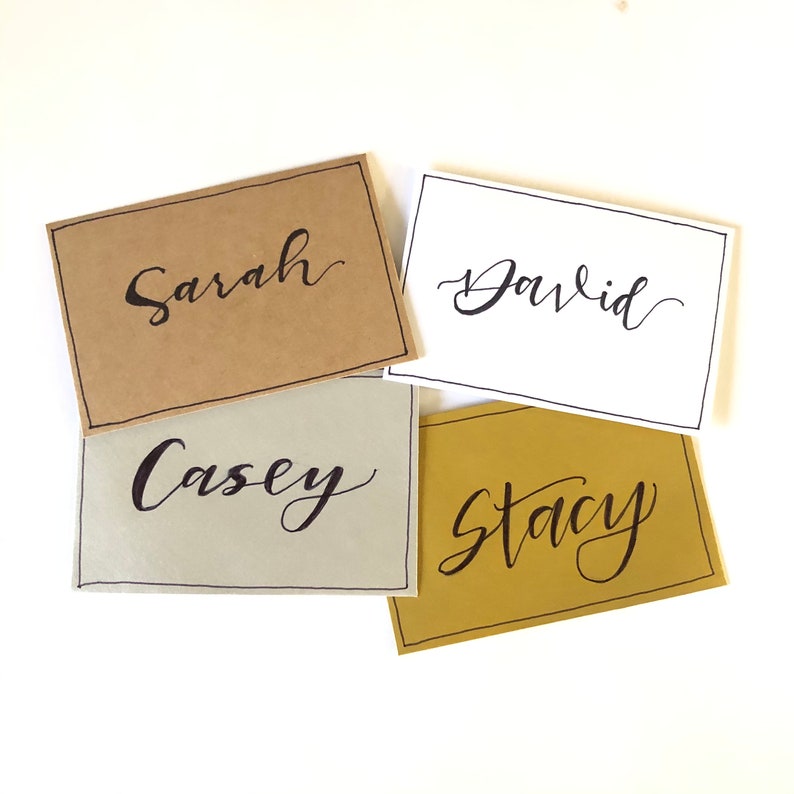 May include: Four place cards with names written in calligraphy. The cards are brown, white, grey, and gold.