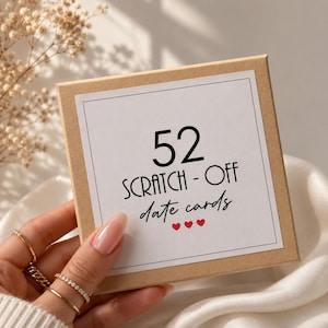 May include: A square kraft paper box with a white label featuring the black text 52 SCRATCH-OFF date cards and three small red hearts. The label is framed by a thin black border, and the box is held against a soft white fabric background.