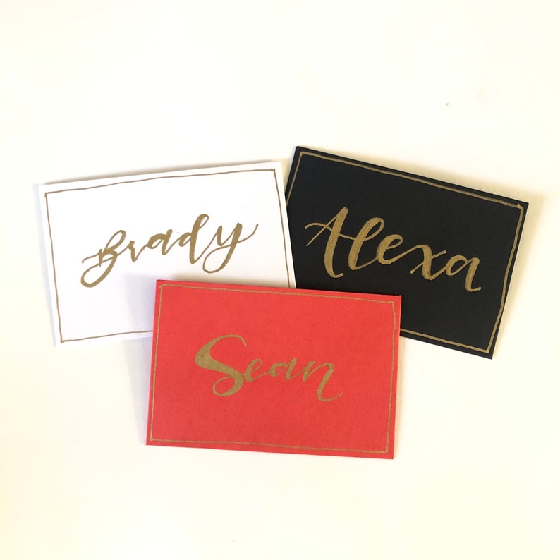May include: Three place cards with gold calligraphy names on different coloured backgrounds. The names are "Brady" on a white background, "Alexa" on a black background, and "Sean" on a red background.