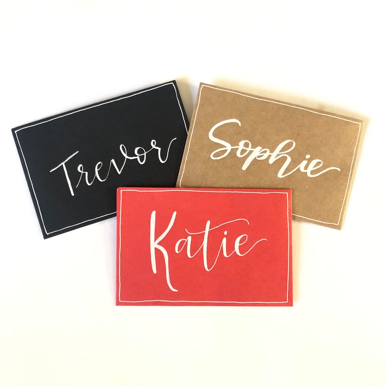 May include: Three place cards with names written in white calligraphy. The cards are on a white background. The names are "Trevor", "Sophie", and "Katie".