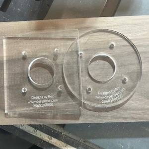 May include: Two clear acrylic router templates, one square and one circular, each with a central hole and mounting holes. The square template has the text "Designs by Rex www.designsbr.com 256010RBSS". The circular template has the text "Designs by Rex www.designsbr.com 256010RBSC".