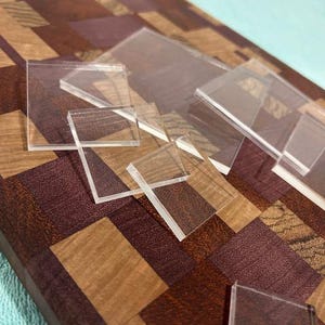 May include: A wooden cutting board with a mosaic pattern of brown, tan, and purple wood pieces. Several clear, square acrylic pieces are scattered across the board, creating a contrast in textures and materials.