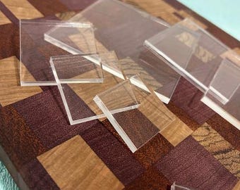 May include: A wooden cutting board with a mosaic pattern of brown, tan, and purple wood pieces. Several clear, square acrylic pieces are scattered across the board, creating a contrast in textures and materials.