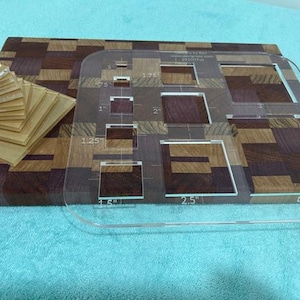 May include: A wooden cutting board with a geometric pattern in shades of brown and purple. A clear acrylic quilting template with various square and rectangular cutouts is on top. A stack of small wooden squares is on the left.