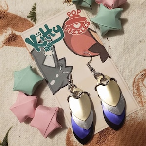 May include: A pair of silver and purple layered dangle earrings with a heart-shaped design. The earrings are hanging from a card with the text "Kitty Pop Fizzles" on it.