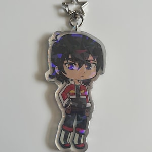 May include: A holographic keychain featuring a chibi-style character with black hair and purple eyes. The character wears a red jacket with black and yellow accents, black pants, and red boots. A silver star-shaped charm is attached to the top.