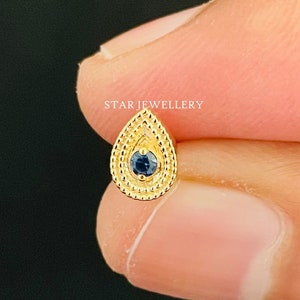 Könnte beinhalten: Ein goldener Nasenpiercing in Tropfenform mit einem blauen Edelstein in der Mitte. Der Tropfen ist von einem Perlmuster umgeben.