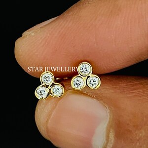 May include: A pair of gold stud earrings, each featuring three round, clear gemstones arranged in a triangular cluster. The earrings are held against a dark background. The text "STAR JEWELLERY" is visible.