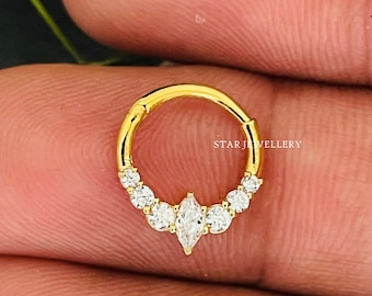 May include: Gold nose ring with a marquise-shaped center stone and smaller round stones around the ring. The ring is on a finger.