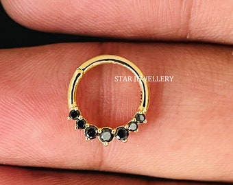 Puede incluir: Anillo de nariz de oro con una fila de piedras preciosas negras. El anillo está abierto y tiene un cierre de bisagra. "STAR JEWELLERY" está impreso en el anillo.