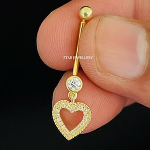 Op de afbeelding: Gouden navelring met een heldere kristal en een hangende hartvormige bedel. De hartvormige bedel is versierd met kleinere heldere kristallen.