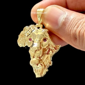 May include: Gold pendant featuring a detailed lion head with red gemstone eyes. The lion's head is sculpted in a 3D style and appears to be part of a larger design.