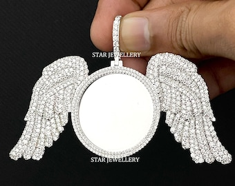 Puede incluir: Un colgante de plata con un inserto de foto redondo y transparente rodeado por un halo de piedras blancas brillantes. Dos alas, también cubiertas de piedras blancas, se extienden desde los lados del colgante. El colgante está etiquetado como "STAR JEWELLERY".
