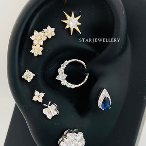 May include: A variety of ear piercings with different styles and gemstones, including a star, a flower, a butterfly, and a teardrop with a blue sapphire. The text "STAR JEWELLERY" is visible in the image.