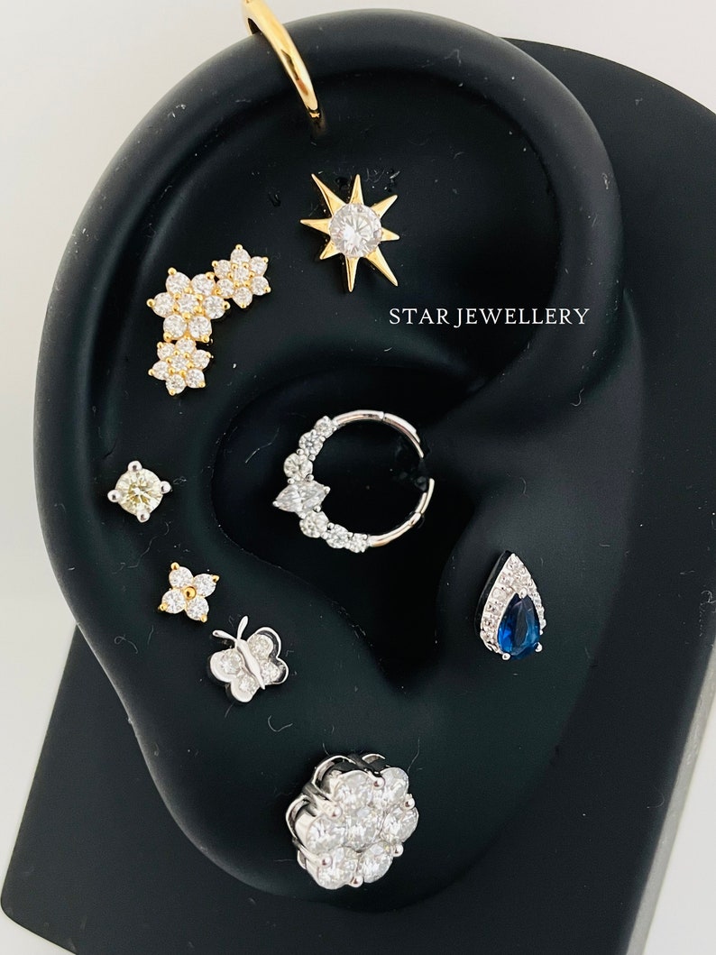 May include: A variety of ear piercings with different styles and gemstones, including a star, a flower, a butterfly, and a teardrop with a blue sapphire. The text "STAR JEWELLERY" is visible in the image.