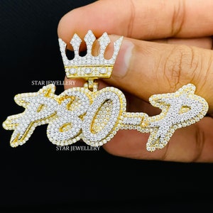 May include: A gold pendant with a crown on top and the numbers "7807" set with diamonds. The pendant is held in a hand.
