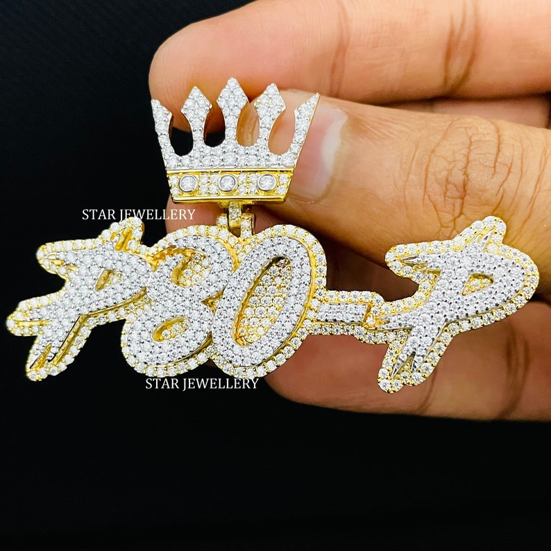 May include: A gold pendant with a crown on top and the numbers "7807" set with diamonds. The pendant is held in a hand.