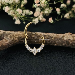 May include: Gold nose ring with a marquise-shaped center stone and smaller round stones on the sides. The ring is on a wooden surface.