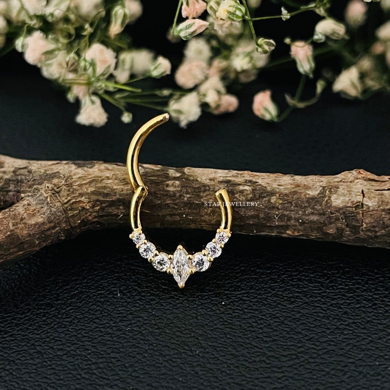 May include: Gold nose ring with a marquise-shaped center stone and smaller round stones on the sides. The ring is on a wooden surface.