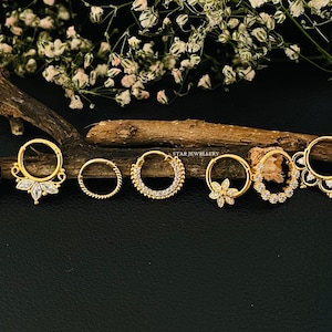 May include: Seven gold nose rings with clear gemstones. The rings are arranged in a row on a wooden surface. The rings have different designs, including a simple circle, a circle with a row of gemstones, and a circle with a floral design.