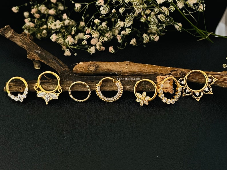 May include: Seven gold nose rings with clear gemstones. The rings are arranged in a row on a wooden surface. The rings have different designs, including a simple circle, a circle with a row of gemstones, and a circle with a floral design.